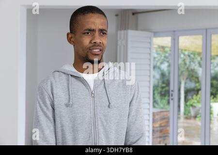 Afroamerikaner, der in der Nähe von Glasschiebetüren im Wohnzimmer steht und einen grauen Hoodie trägt, der reagiert Stockfoto