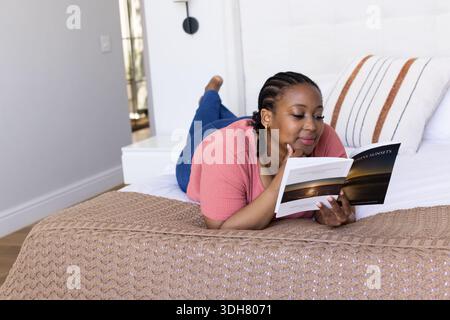Afroamerikanerin, die auf dem Magen liegt und auf dem Bett einen Sunset-Taschenbuch liest, mit rosa Oberteil und Jeans Stockfoto