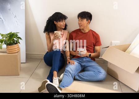 Verschiedene Paare sitzen auf gefliestem Boden mit Keramikbechern, in Jeans und T-Shirts, packen Kartons aus Stockfoto