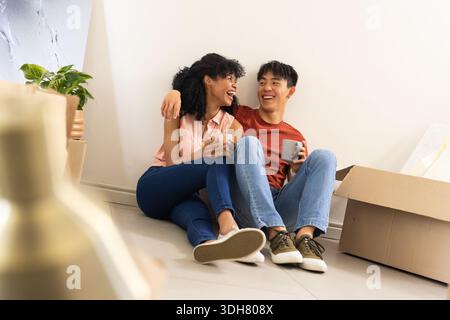 Verschiedene Paare sitzen zu Hause auf dem Boden und lachen, während sie Keramikbecher in der Nähe von gestapelten Kisten halten Stockfoto