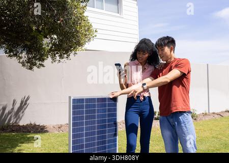 Diverse Paare stehen auf dem Rasen neben Solarpanel, Frau mit Smartphone, Mann zeigt Armbanduhr Stockfoto