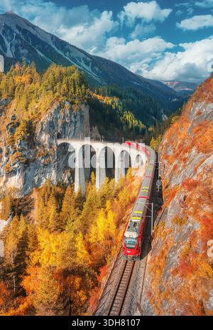 Aus der Vogelperspektive der modernen roten Bahn auf dem Landwasserviadukt in alpinen Bergen, farbenfroher Wald bei Sonnenuntergang im Herbst. Bernina Express, Schweiz. Draufsicht Stockfoto