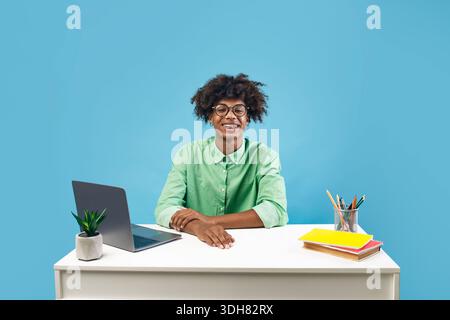 Porträt eines glücklichen afroamerikanischen Mannes, der mit Büchern und Laptop am Schreibtisch sitzt und auf blauem Hintergrund die Kamera anlächelt Stockfoto