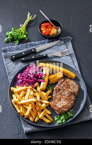 Mittelseltenes Rindfleischsteak im texanischen Stil, serviert mit Pommes frites, Rotkohl-Salat und gegrillten Maisrippchen auf einem Teller auf Betontisch mit Besteck, V Stockfoto