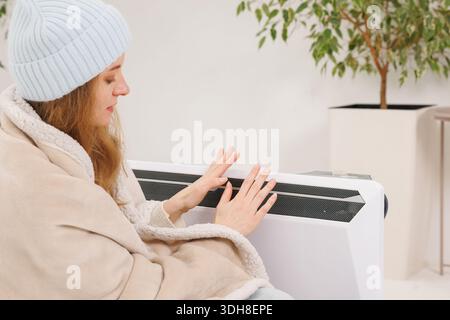 Nahaufnahme einer Frau in Beanie und Decke, die die Hände durch eine tragbare elektrische Heizung in einer kalten Wohnung während des Winters energiesparend wärmt Stockfoto