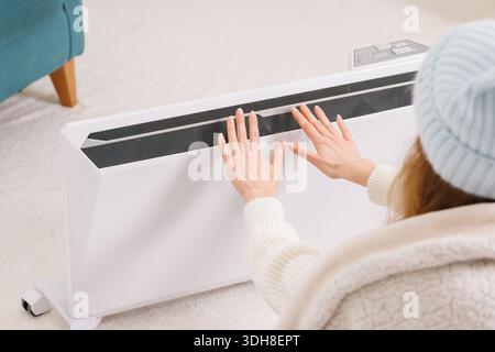 Nahaufnahme einer Frau, die während des Winters die Hände durch eine tragbare elektrische Heizung in einem kalten, minimalistischen Wohnraum wärmt und sich auf Energiesparen und Komfort konzentriert Stockfoto