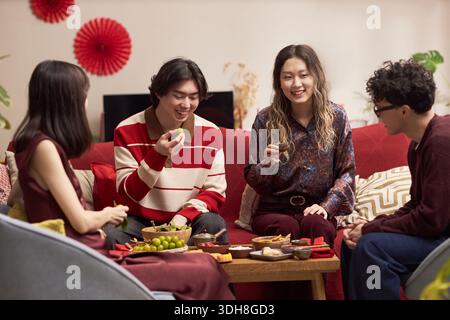 Gruppe junger erwachsener asiatischer Männer und Frauen, die zusammen auf dem Sofa sitzen und während der Tet-Feier traditionelle Weihnachtsessen und -Getränke genießen, lächeln und miteinander interagieren Stockfoto