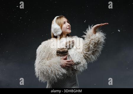 Junge Frau in flauschigem Mantel und Ohrenschützern, die Schnee im Studio auf dunklem Hintergrund für Winteranzeigen und saisonales Design fangen Stockfoto
