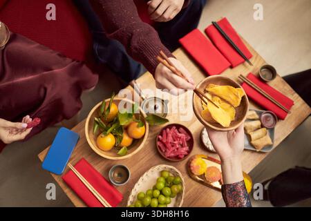 Asiatische junge Erwachsene und Teenager teilen sich traditionelle Feiertagsjause mit Essstäbchen, die Hände reichen über einen Holztisch mit getrockneten Früchten, Zitrusfrüchten, Süßigkeiten und festlichen roten Servietten während der Tet-Feier Stockfoto