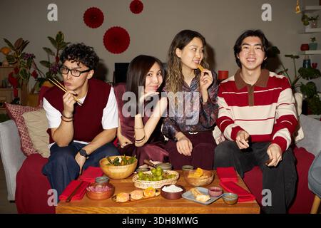 Gruppe asiatischer junger Erwachsener, die auf dem Sofa sitzen und gemeinsam traditionelle Tet-Feiertagsmahlzeiten genießen, lächeln und Essstäbchen, festliche Dekoration und vietnamesische Gerichte auf dem Tisch verwenden Stockfoto