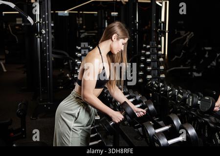 Die junge athletische Frau wählt im Fitnessstudio eine Kurzhantel, die sich auf Krafttraining und Fitness-Workout vorbereitet Stockfoto