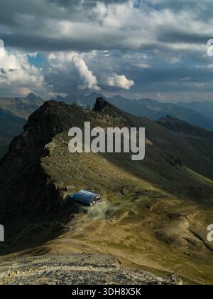 Eine einsame Berghütte befindet sich in der Nähe eines Sattels auf einem zerklüfteten Bergkamm in den europäischen Alpen, während Sonnenstrahlen durch Wolken brechen und gewundene Fußwege erleuchten. Stockfoto