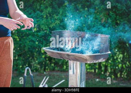 Das Feuer in einem Fleischgrill anzünden, Nahaufnahme eines Grills und die Hände einer Person zünden das Feuer auf einem Grill an Stockfoto