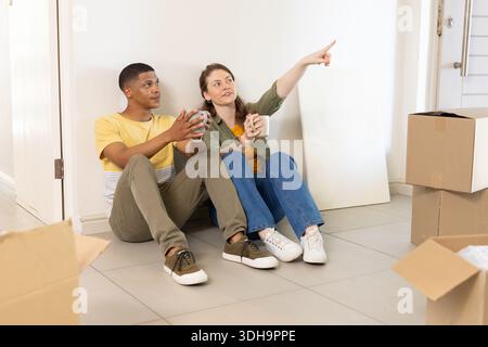 Verschiedene Paare sitzen auf gefliestem Boden am Eingang und halten Tassen und zeigen auf offene Umzugskästen Stockfoto