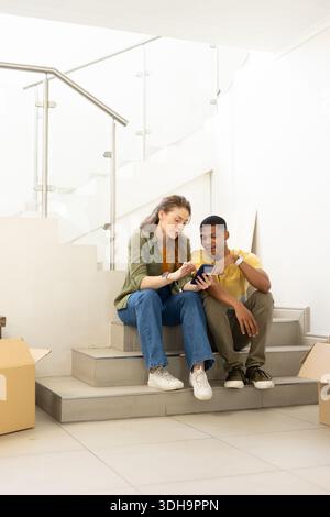 Verschiedene Paare sitzen auf gekachelten Treppen und landen zu Hause, packen Kartons aus und überprüfen das Smartphone Stockfoto