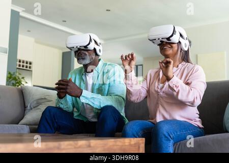 Verschiedene Seniorenpaare tragen weiße VR-Headsets, greifen und lächeln auf dem grauen Sofa am Holztisch Stockfoto