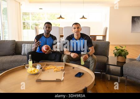 Verschiedene männliche Freunde sitzen auf Sofa im Wohnzimmer, passende Shirts, halten Fußball und Chips Stockfoto