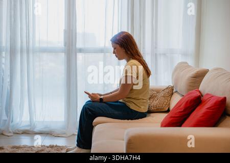 Frau sitzt auf einem Sofa im Profil mit einem Smartphone in der Nähe eines hellen Fensters, gemütlicher Lebensstil zu Hause. Stockfoto