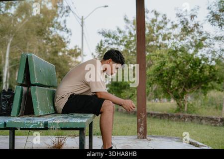 Seitenprofil eines jungen Mannes, der auf einer Parkbank unter einem Pavillon sitzt, nachdenkliche und ruhige Stimmung. Stockfoto