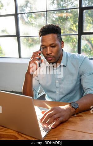 Ein Erwachsener Afroamerikaner trägt ein blaues Hemd, eine Uhr, tippt auf einem Laptop am Holztisch, hält das Telefon Stockfoto