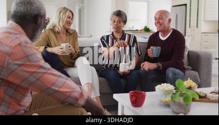 Sprechende Gruppe von vier Erwachsenen, die an einem weißen Couchtisch im Wohnzimmer sitzen und Keramikbecher halten Stockfoto