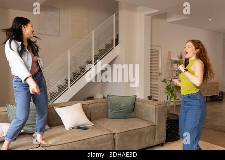 Verschiedene Freundinnen tanzen und singen auf einem Sofa im Wohnzimmer mit Smartphone Stockfoto