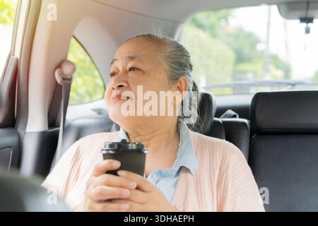 Glückliche ältere asiatische Frau sitzt auf dem Beifahrersitz mit einer Tasse Tee im Auto und genießt die Fahrt Stockfoto