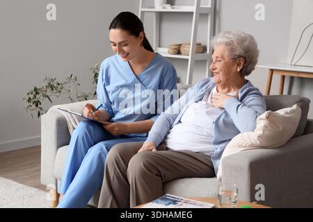 Ältere Frau mit Physiotherapeutin, die zu Hause auf dem Sofa in die Zwischenablage schreibt Stockfoto