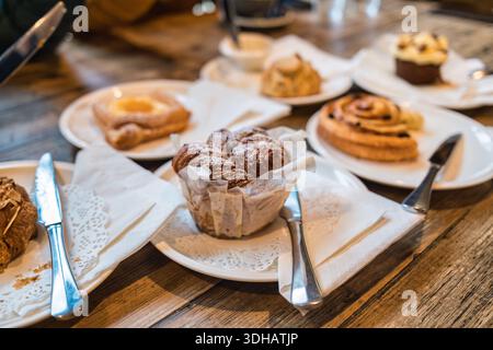 Verschiedene Gebäcksorten, Muffins, Zimtrollen, Scone und Gebäck auf Holztisch, Teller mit Besteck in der Bäckerei Stockfoto