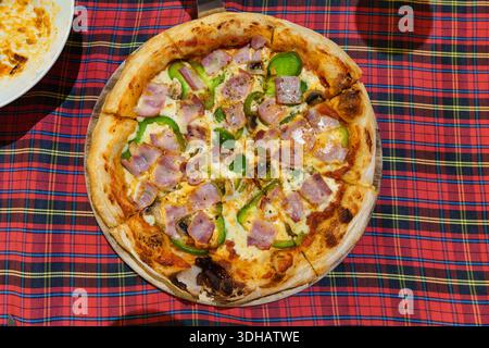 Auffällige gebackene Pizza mit Schinken, grünem Paprika und gewürztem Käse, serviert auf Teller auf Tartantischdecke Stockfoto