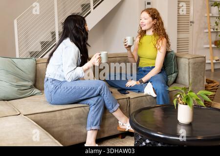 Verschiedene Freundinnen tragen eine Jeansjacke in Limettengrün, trinken Tassen und plaudern auf einem Sofa Stockfoto