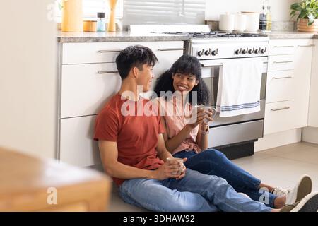 Diverse Jeans sitzen auf gefliestem Küchenboden und halten Keramikbecher in der Nähe des Ofens Stockfoto
