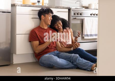 Verschiedene Paare sitzen auf gefliestem Küchenboden und lehnen sich an die unteren Schränke und halten Keramikbecher Stockfoto