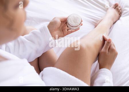 Hände halten das Glas mit Creme offen, verteilen Lotion auf Schienbein, während sie weißen Bademantel auf dem Bett tragen Stockfoto