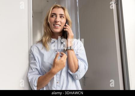 MittelErwachsene Frau, die sich in der Tür beugt, gestreiftes Hemd, Uhr und Armband trägt und telefoniert Stockfoto