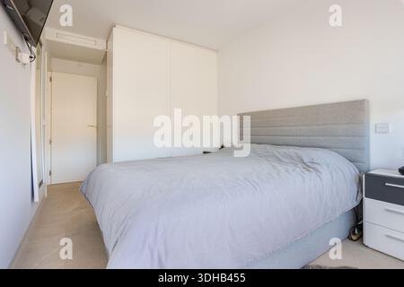 Helles Schlafzimmer mit weichen grauen Betten, eingebautem Kleiderschrank und modernem Fernseher an der Wand Stockfoto