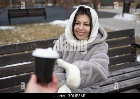 Eine Frau in Pelzmantel und Kopftuch sitzt auf einer Parkbank und lächelt, während jemand ohne Kamera ihr eine Tasse Kaffee an einem kalten Wintertag anbietet Stockfoto