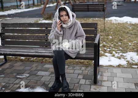 Eine Frau mit Pelzmantel, weißen Handschuhen und Kopftuch sitzt im Winter auf einer Holzbank in einem Park. Sie spricht mit einem ernsten Ausdruck auf ihrem Handy. Stockfoto