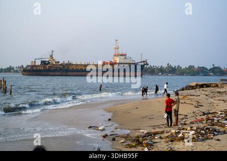Fort Kochi, Indien - 16. März 2023: Mehrere Menschen stehen und gehen an einem Sandstrand entlang, der mit Trümmern und Müll verstreut ist, während ein großes Frachtschiff vor der Küste vor Anker liegt. Stockfoto