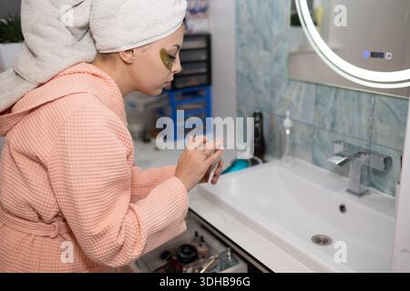 Eine junge Frau mit einem Handtuch auf dem Kopf trägt Gesichtscreme in ihrem modernen Badezimmer auf. Sie trägt Augenflecken und einen rosa Mantel, während sie am Waschbecken steht. Morgenroutine. Stockfoto