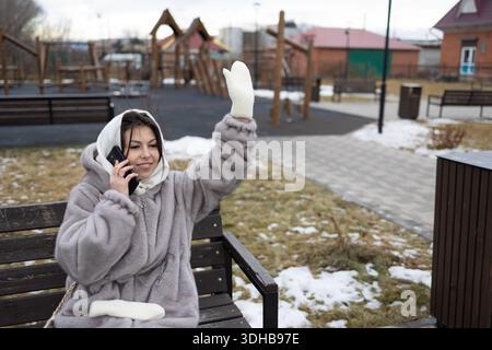 Eine Frau sitzt auf einer Bank in einem Park mit Mantel und Handschuhen. Sie hält ein Telefon am Ohr und winkt mit der anderen Hand. Schnee liegt auf dem Boden und Spielplatzstrukturen sind hinter ihr sichtbar. Stockfoto