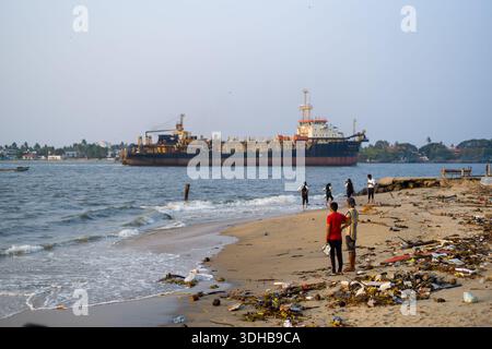 Fort Kochi, Indien - 16. März 2023: Mehrere Menschen stehen und gehen an einem Sandstrand entlang, der mit Trümmern und Müll verstreut ist, während ein großes Frachtschiff vor der Küste vor Anker liegt. Stockfoto