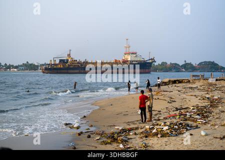 Fort Kochi, Indien - 16. März 2023: Mehrere Menschen stehen und gehen an einem Sandstrand entlang, der mit Trümmern und Müll verstreut ist, während ein großes Frachtschiff vor der Küste vor Anker liegt. Stockfoto
