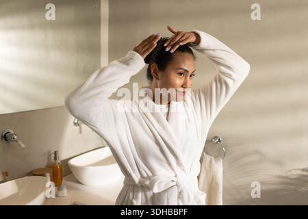 Asiatische Frau, die im Badezimmer am Eitelbereich steht, in weißem Bademantel, die Haare durch den Spiegel in Brötchen einstellt Stockfoto