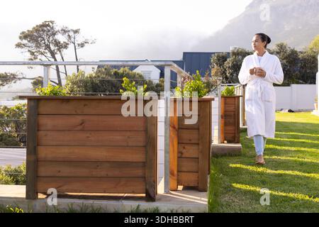 Asiatische Frau im Gewand, die auf der Terrasse neben Pflanzgefäßen steht und einen Becher hält, der nach rechts schaut, Kopierraum Stockfoto