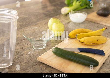 Hölzerne Schneidebrett auf Granittheke mit Gurke und blauem Messer, Bananen, Schüssel Stockfoto