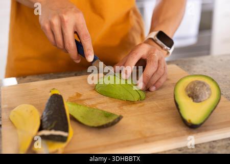 Geschälte Avocado wird auf hölzernem Schneidebrett auf gesprenkelter Arbeitsplatte in der Nähe der Avocadohälfte geschnitten Stockfoto