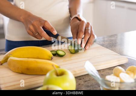 Gurke wird auf hölzernem Schneidebrett auf Steinplatte geschnitten, Bananen und Schüssel sichtbar Stockfoto