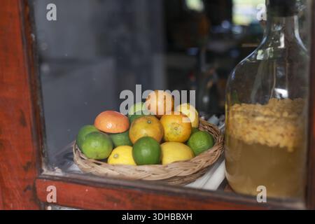 Frischer Korb gefüllt mit Zitrusfrüchten auf der Fensterbank neben einem Glasgefäß mit konserviertem flüssigem Getränk mit warmer, gemütlicher Stimmung hausgemachter Presse Stockfoto