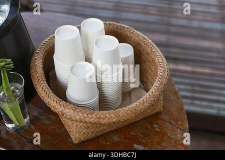 Stapel weißer Einweg-Pappbecher in rundem braunem, gewebtem Korb auf Holztisch, bereit für den Service mit natürlicher rustikaler Atmosphäre Stockfoto